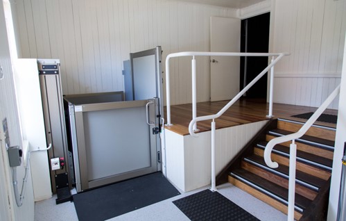 A platform lift, which has waist high doors and open space above it sits next to a flight of stairs